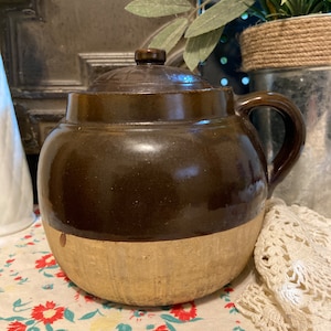 May include: A brown glazed crock pot with a lid and a handle. The crock pot has a light brown band around the bottom.