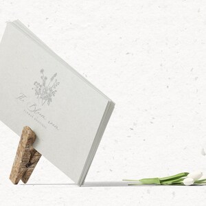 May include: A white card with the text "The Bloom Room Flower Boutique" and a floral illustration is held up by a wooden clothespin. A bouquet of white tulips lies on a white textured surface.