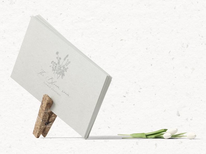 May include: A white card with the text "The Bloom Room Flower Boutique" and a floral illustration is held up by a wooden clothespin. A bouquet of white tulips lies on a white textured surface.