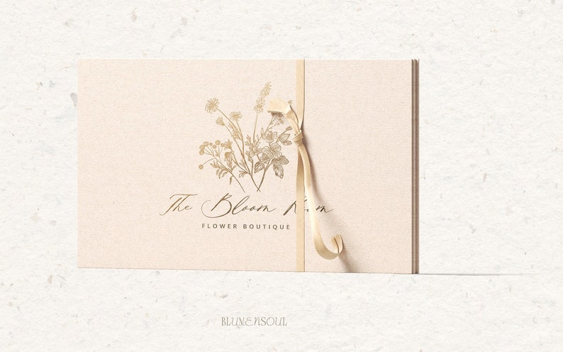 May include: A beige card with a gold ribbon tied around it. The card has a floral design and the text "The Bloom Room Flower Boutique".