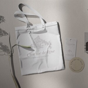 May include: A white tote bag with the text "Charme de Rose Vintage Clothing Est. 2021" and a floral design. The bag is on a light gray surface with a brown tag and a small concrete block with a leaf.