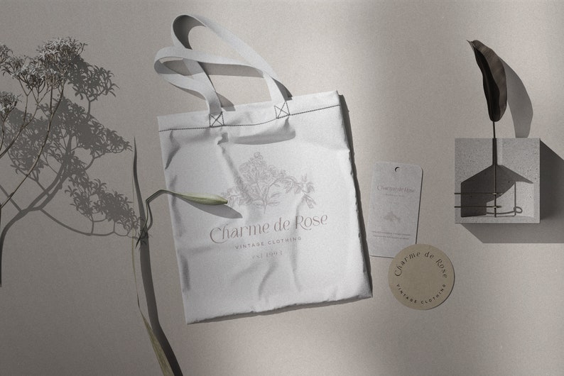 May include: A white tote bag with the text "Charme de Rose Vintage Clothing Est. 2021" and a floral design. The bag is on a light gray surface with a brown tag and a small concrete block with a leaf.