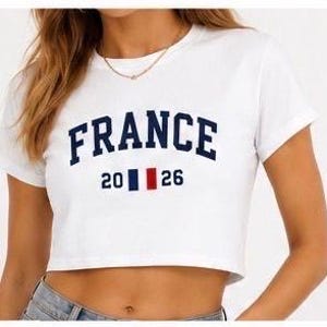 Puede incluir: Top corto blanco con la palabra "FRANCE" en letras arqueadas de color azul marino. Debajo de la palabra "FRANCE" est&aacute; el n&uacute;mero "2026" con un dise&ntilde;o de rayas verticales azul marino, blanco y rojo. La camiseta tiene mangas cortas.