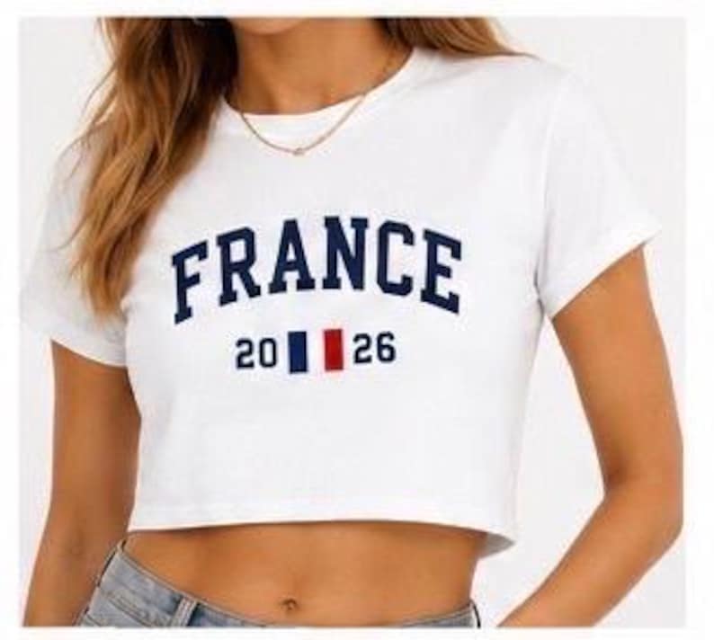 Puede incluir: Top corto blanco con la palabra "FRANCE" en letras arqueadas de color azul marino. Debajo de la palabra "FRANCE" est&aacute; el n&uacute;mero "2026" con un dise&ntilde;o de rayas verticales azul marino, blanco y rojo. La camiseta tiene mangas cortas.