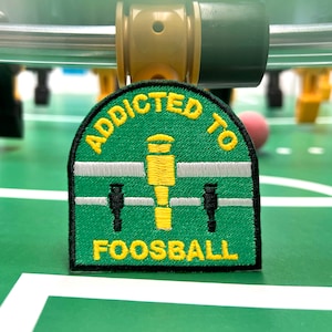Puede incluir: Parche bordado verde y amarillo con el texto "Addicted to Foosball" y un gráfico de un jugador de futbolín.