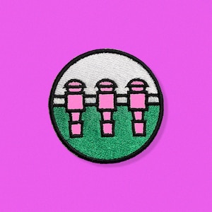 May include: A round embroidered patch featuring three pink figures standing in a row on a green field. The figures have white hats and black outlines. The field is surrounded by a white border and a black outline.