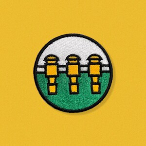 May include: A green and white embroidered patch with three yellow figures standing in a row. The figures have black outlines and white accents. The patch is circular and has a black border.