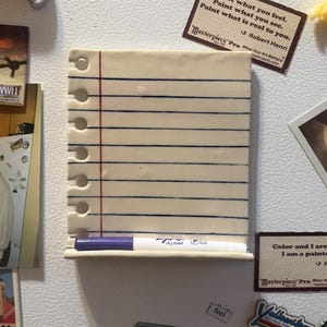 May include: A rectangular ceramic dry erase board shaped like a piece of notebook paper with horizontal blue lines and a vertical red margin line. The board features a row of circular cutouts along the left side and a small ledge holding a purple marker.