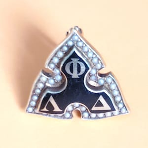 May include: A silver triangular pin with a black enamel center. The pin is decorated with white pearls around the edges and features the Greek letters "ΦΔΔ" in gold.