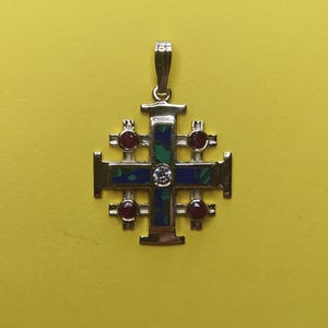 May include: A gold-colored pendant in the shape of a cross, featuring a central diamond and four smaller red gemstones. The cross has a blue and green patterned inlay. The pendant hangs from a small loop.