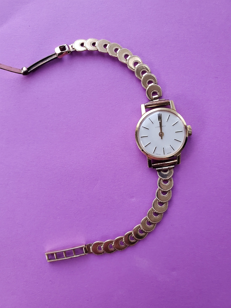 K&ouml;nnte beinhalten: Eine goldene Armbanduhr mit wei&szlig;em Zifferblatt und einem goldenen Gliederarmband. Das Zifferblatt der Uhr hat schwarze r&ouml;mische Ziffern als Stundenmarkierungen und schwarze Zeiger. Die Uhr befindet sich auf einem violetten Hintergrund.