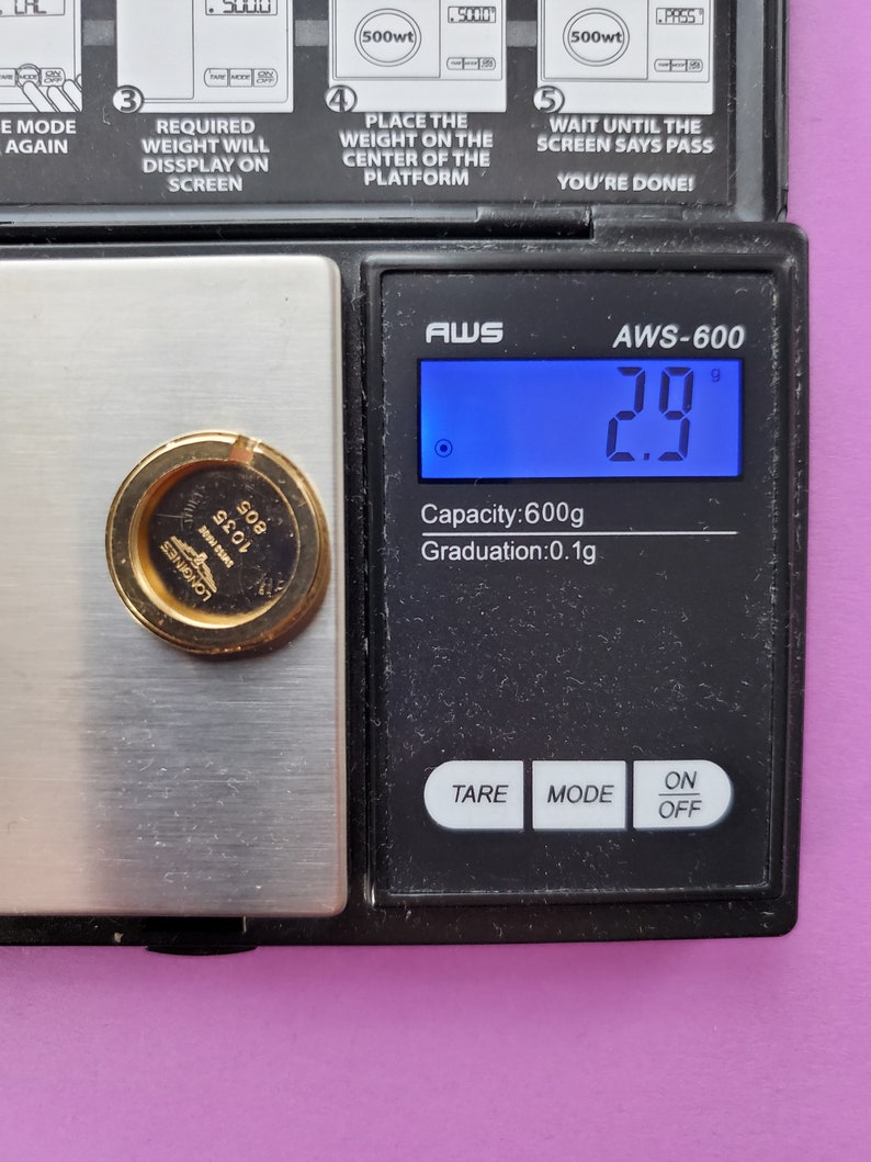 K&ouml;nnte beinhalten: Eine digitale Waage mit einem goldfarbenen Objekt auf der Plattform. Die Waage zeigt 2,9 Gramm an. Die Waage hat ein schwarzes Display mit wei&szlig;em Text, auf dem "AWS-600" und "Kapazit&auml;t: 600 g Aufl&ouml;sung: 0,1 g" steht. Die Waage hat drei Tasten mit den Beschriftungen "TARE", "MODE" und "EIN/AUS".
