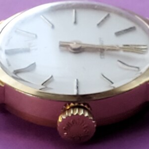 K&ouml;nnte beinhalten: Eine goldfarbene Armbanduhr mit wei&szlig;em Zifferblatt und r&ouml;mischen Ziffern f&uuml;r die Stunden. Die Uhr hat ein goldfarbenes Armband und ein rundes Geh&auml;use.