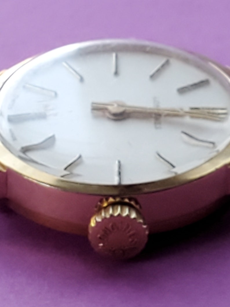 K&ouml;nnte beinhalten: Eine goldfarbene Armbanduhr mit wei&szlig;em Zifferblatt und r&ouml;mischen Ziffern f&uuml;r die Stunden. Die Uhr hat ein goldfarbenes Armband und ein rundes Geh&auml;use.