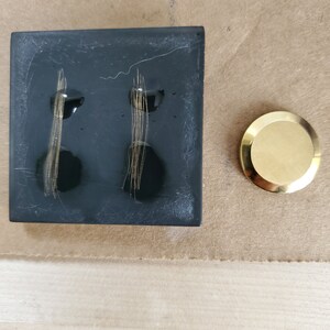 K&ouml;nnte beinhalten: Eine schwarze quadratische Fliese mit zwei goldenen Linien darauf. Die Fliese liegt auf einer braunen Oberfl&auml;che neben einer goldenen runden Scheibe.