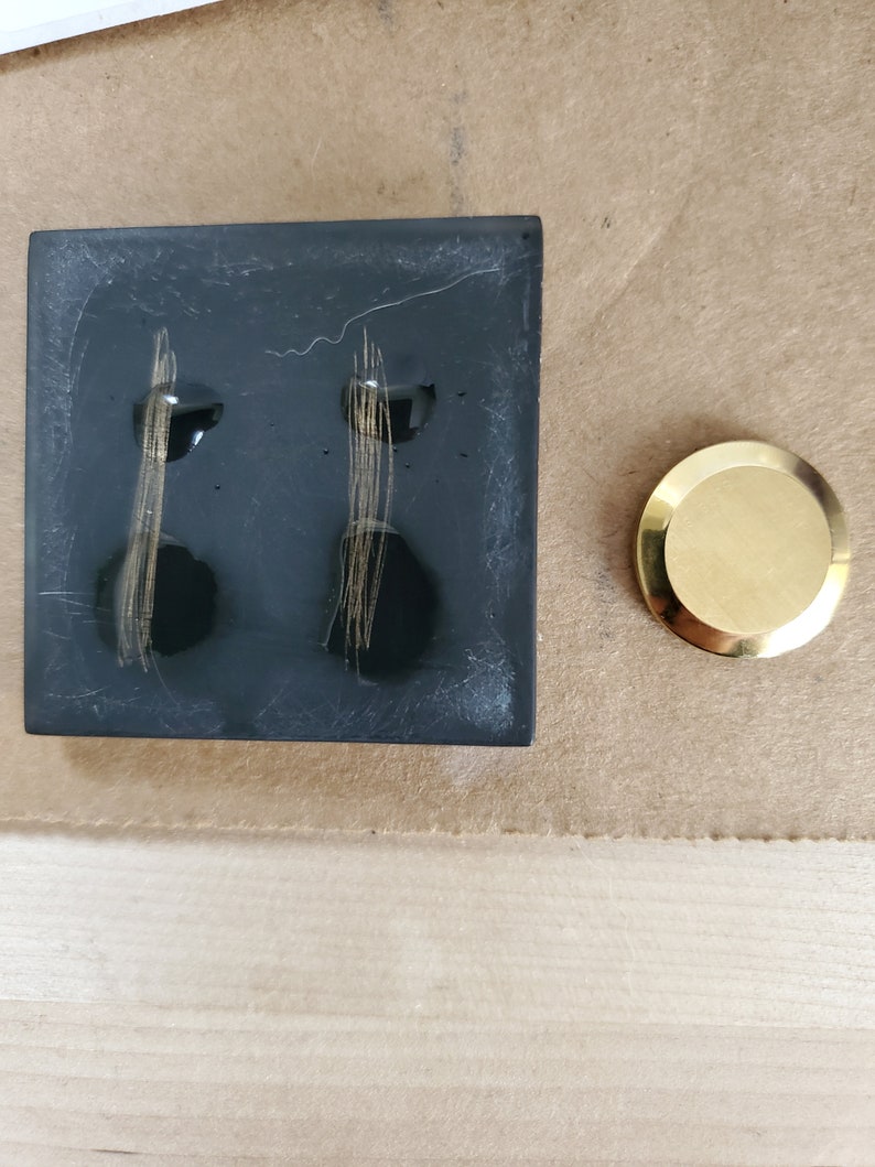 K&ouml;nnte beinhalten: Eine schwarze quadratische Fliese mit zwei goldenen Linien darauf. Die Fliese liegt auf einer braunen Oberfl&auml;che neben einer goldenen runden Scheibe.
