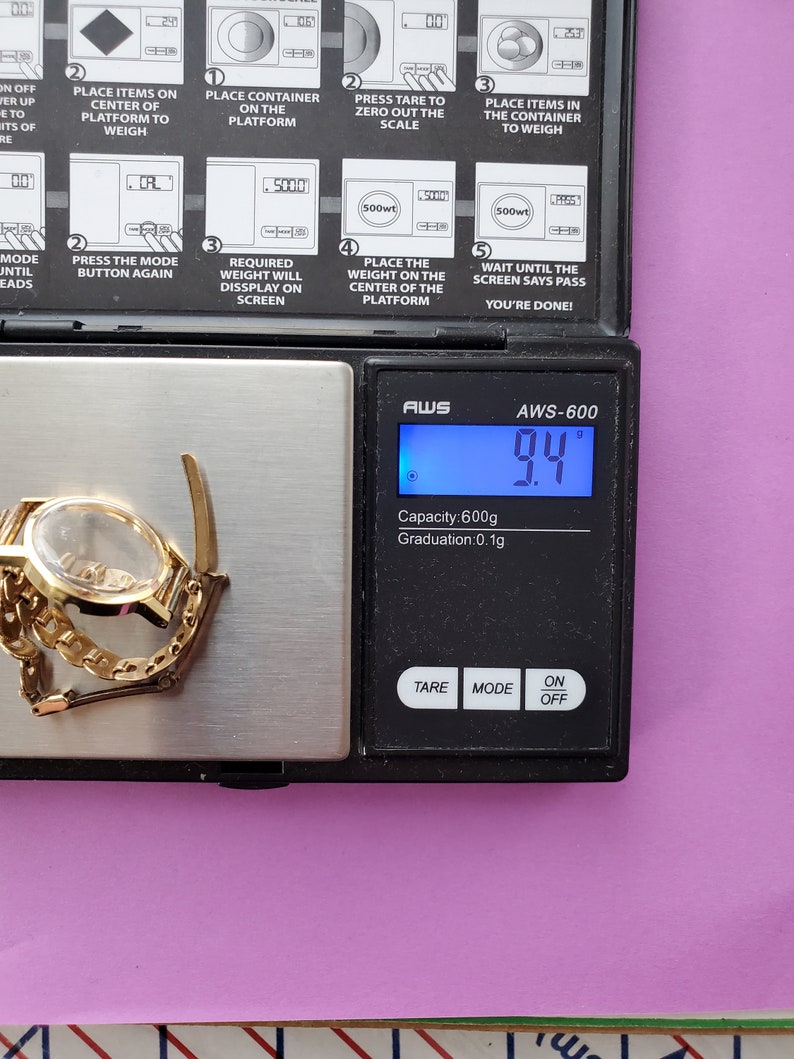K&ouml;nnte beinhalten: Eine schwarze digitale Waage mit einer silbernen Plattform. Die Waage zeigt 9,4 Gramm an. Die Waage hat eine Kapazit&auml;t von 600 Gramm und eine Abstufung von 0,1 Gramm. Die Waage verf&uuml;gt &uuml;ber Tasten f&uuml;r Tara, Modus und Ein/Aus. Die Waage befindet sich auf einer rosa Oberfl&auml;che. Eine goldene Uhr befindet sich auf der Plattform.