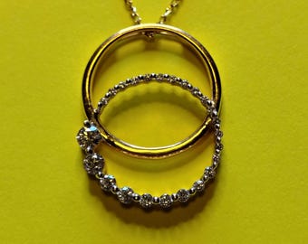 Könnte beinhalten: Eine goldene Halskette mit einem Anhänger aus zwei ineinandergreifenden Kreisen. Ein Kreis ist ein schlichter, polierter Goldring, während der andere ein silberfarbener Ring ist, der mit einer Reihe kleiner, klarer, runder Steine besetzt ist.