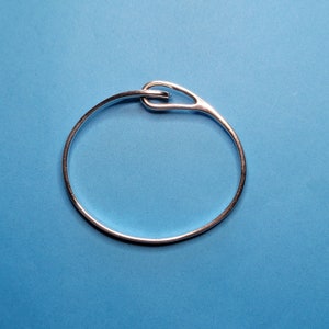 May include: A silver bangle bracelet with a simple, modern design. The bracelet is made of a single piece of metal that is bent into a circle and then twisted at the clasp.