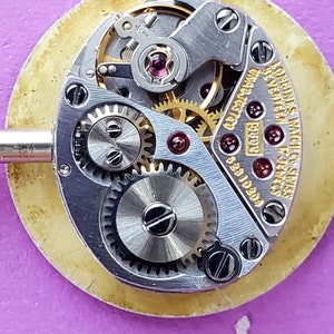 K&ouml;nnte beinhalten: Nahaufnahme eines silbernen Uhrwerks mit dem sichtbaren Text "Longines Watch Co. Swiss" und "17 Jewels". Das Uhrwerk hat einen goldfarbenen Rotor und mehrere Zahnr&auml;der.