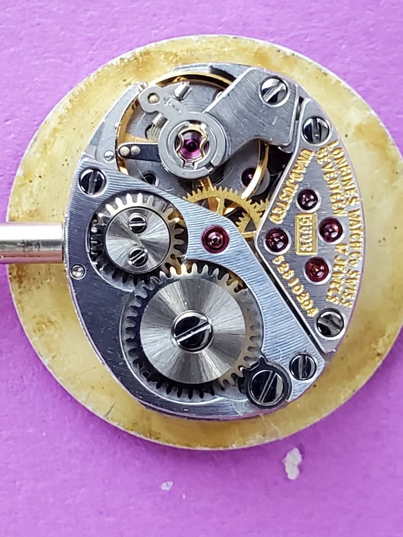 K&ouml;nnte beinhalten: Nahaufnahme eines silbernen Uhrwerks mit dem sichtbaren Text "Longines Watch Co. Swiss" und "17 Jewels". Das Uhrwerk hat einen goldfarbenen Rotor und mehrere Zahnr&auml;der.