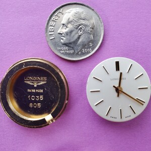 K&ouml;nnte beinhalten: Nahaufnahme von drei Uhrenteilen: ein goldfarbener Uhrenboden mit dem Text "Longines Swiss Made 1035 805", ein wei&szlig;es Zifferblatt mit schwarzen Zeigern und dem Text "Longines" und eine silberfarbene US-Dime-M&uuml;nze mit dem Text "Liberty In God We Trust 2016 D".