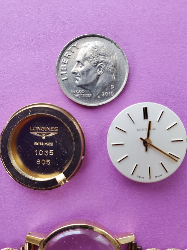 K&ouml;nnte beinhalten: Nahaufnahme von drei Uhrenteilen: ein goldfarbener Uhrenboden mit dem Text "Longines Swiss Made 1035 805", ein wei&szlig;es Zifferblatt mit schwarzen Zeigern und dem Text "Longines" und eine silberfarbene US-Dime-M&uuml;nze mit dem Text "Liberty In God We Trust 2016 D".