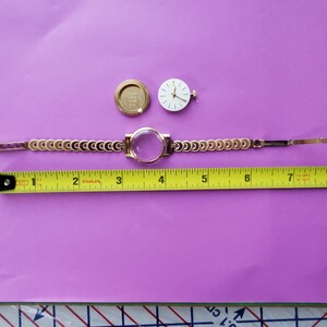 K&ouml;nnte beinhalten: Eine goldfarbene Uhr mit wei&szlig;em Zifferblatt und schwarzen Zeigern. Die Uhr ist zerlegt und auf einer violetten Oberfl&auml;che mit einem Ma&szlig;band ausgelegt. Das Zifferblatt der Uhr hat einen Durchmesser von 3,8 cm.