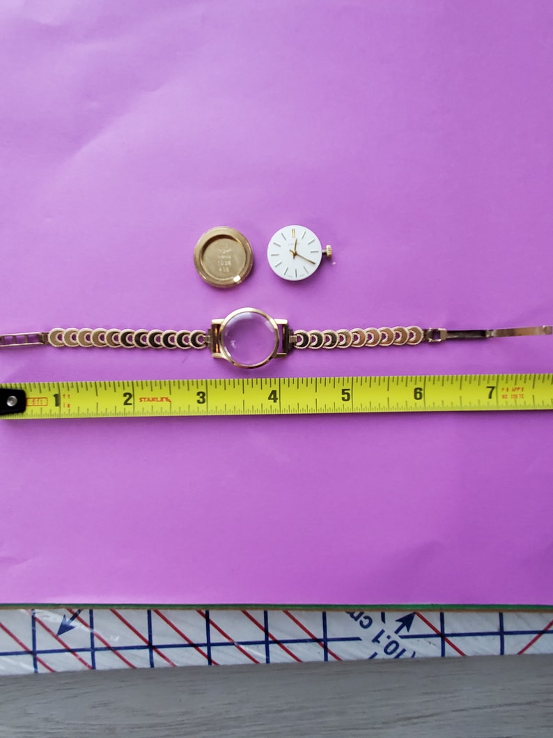 K&ouml;nnte beinhalten: Eine goldfarbene Uhr mit wei&szlig;em Zifferblatt und schwarzen Zeigern. Die Uhr ist zerlegt und auf einer violetten Oberfl&auml;che mit einem Ma&szlig;band ausgelegt. Das Zifferblatt der Uhr hat einen Durchmesser von 3,8 cm.