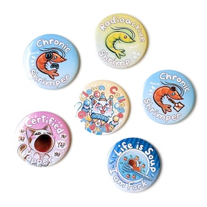 May include: A collection of six colorful round buttons with various designs and text. The buttons feature illustrations of a shrimp, a cat, and a plate of soup. Text includes "Chronic Shrimper," "Radioactive Shrimp," and "Life is Soup I Am Fork."