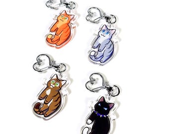 May include: Four different cat keyrings with heart-shaped rings. The cats are orange, grey, brown, and black. Each cat is in a sitting position with its tail curled around its body.