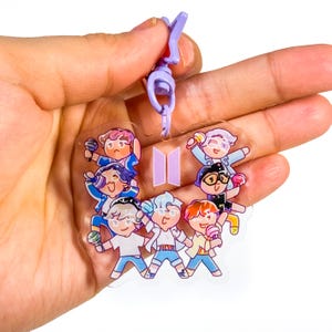 May include: A clear acrylic charm featuring a group of cartoon characters in various outfits, holding microphones and ice cream cones. The charm has a purple clasp and a purple logo. The characters are in shades of blue, pink, orange, and white.