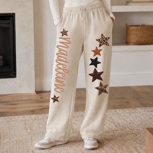 May include: Beige sweatpants featuring a drawstring waist and side pockets. The left leg displays the name "Madeleine" in a cursive script alongside a small star, while the right leg features a vertical arrangement of five stars in various colors and patterns, including leopard print, solid brown, navy blue, and dark brown.