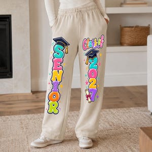 May include: Beige sweatpants featuring a colorful graphic print on the legs. The left leg displays the word SENIOR in large, multi-colored bubble letters topped with a graduation cap. The right leg reads Class of 2027 with a cap and diploma.