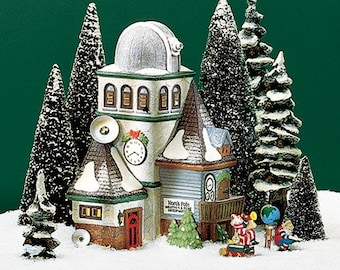 May include: A miniature Christmas village scene with a white building with a green roof and a clock tower. The building has a sign that reads "North Pole Weather & Time Observatory". There are several snow-covered pine trees and a red sled with two figures in it.