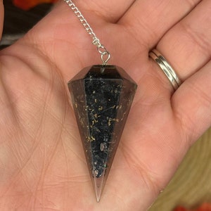 May include: A black and gold crystal pendulum with a silver chain. The pendulum is in the shape of a point and has a dark, almost black, crystal with gold flecks throughout.