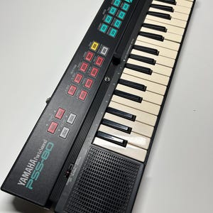 May include: Black Yamaha PortaSound PSS-80 keyboard with white and black keys. The keyboard has various function buttons in red, yellow, and teal. The speaker is located on the bottom right.