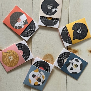 May include: A selection of miniature vinyl records in various coloured sleeves. Each sleeve displays a different cat illustration with the text "King Meow". The records are black with a circular label.
