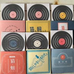 May include: A collection of square cards with a vinyl record design and coloured labels. The cards are arranged with colourful packaging, some with Japanese text. The theme is stationery or gift items.