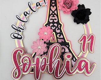 May include: A pink and gold cake topper featuring a black silhouette of the Eiffel Tower, pink flowers, and the text "Oh la la!" and "Sophia 1".