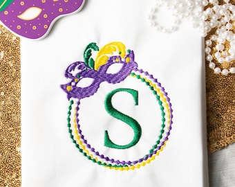 May include: White embroidered kitchen towel with a purple, green, and gold Mardi Gras mask and beads surrounding the letter "S".