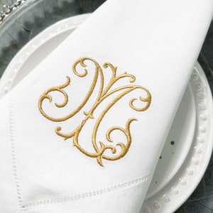 May include: A white linen napkin with a gold embroidered monogram of the letter "M" in a decorative script font.