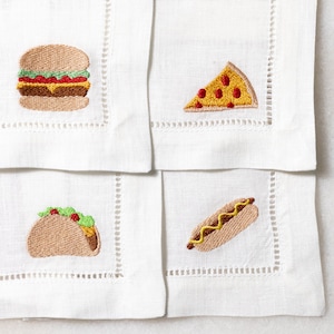 May include: Four white linen napkins with embroidered images of food. The top left napkin has a hamburger, the top right has a slice of pizza, the bottom left has a taco, and the bottom right has a hot dog.