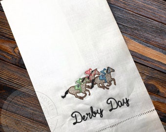 May include: White linen tea towel with embroidered horses and jockeys racing. The text "Derby Day" is embroidered below the horses.