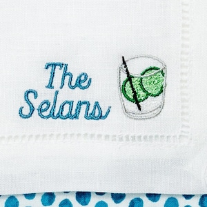 May include: White linen napkin with blue embroidery that reads "The Selans" and a small embroidered image of a glass with a lime wedge and a straw.