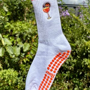 May include: White crew sock with an orange cuff and orange non-slip dots on the sole. A cocktail graphic is on the side. The word "Hour" is printed on the sole.