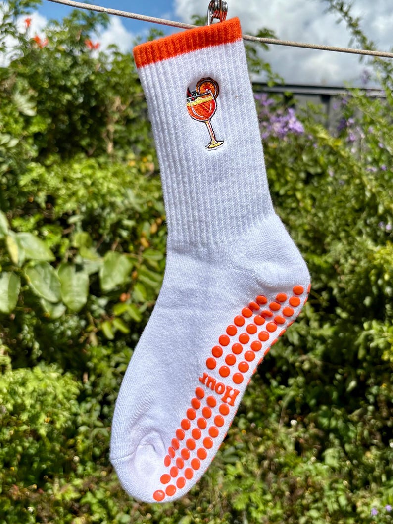 May include: White crew sock with an orange cuff and orange non-slip dots on the sole. A cocktail graphic is on the side. The word "Hour" is printed on the sole.