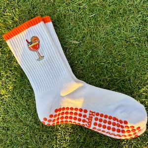 May include: White socks with orange accents and a cocktail embroidery. The socks have orange non-slip grips on the soles and a ribbed cuff. The cocktail design features a red straw and orange slice.