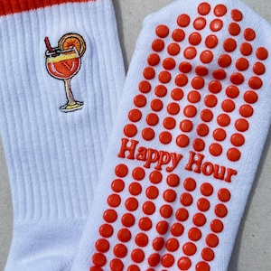 May include: White socks with orange accents and a cocktail embroidery. The socks have orange non-slip grips on the soles and the words "Happy Hour" in orange. The top of the socks are orange.