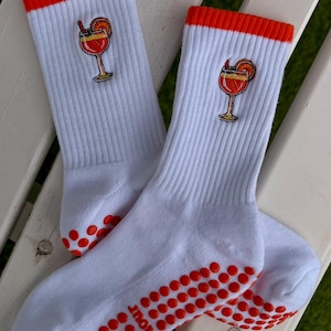 May include: White socks with orange accents and a cocktail embroidery. The socks have a ribbed texture, orange cuffs, and orange non-slip dots on the soles. The cocktail embroidery features a red drink with an orange slice and a straw.